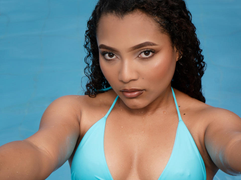 LoraineeRios with curly black hair, wearing a wet, turquoise bikini top, showing her small, round boobs, and gazing at the camera with a serious expression. Blue background.