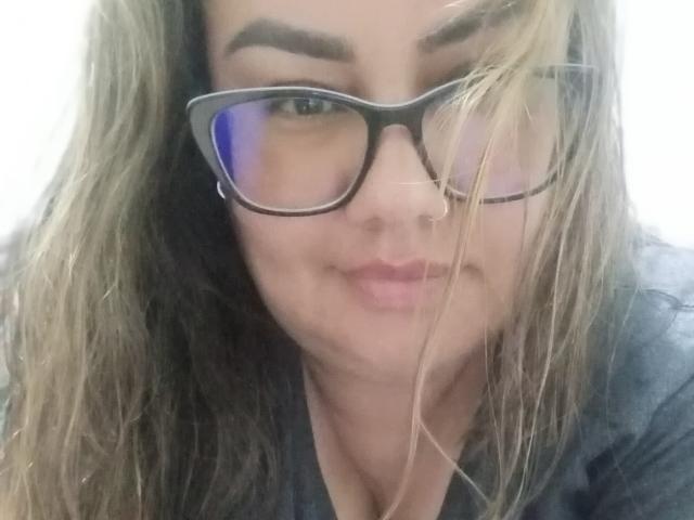 FoxyBell, avec ses cheveux blonds et bruns, porte des lunettes noires, a un léger sourire et regarde l'appareil photo avec les sourcils légèrement relevés. Ses seins sont partiellement visibles..