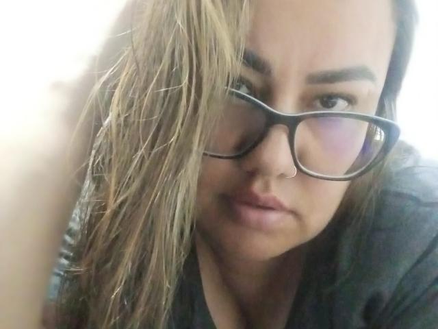 FoxyBell, avec ses cheveux bruns mouillés, ses lunettes noires et son anneau de nez, a l'air légèrement agacée. Ses lèvres boudeuses sont légèrement entrouvertes. Elle porte un t-shirt noir..