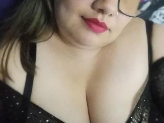 FoxyBell, avec du rouge à lèvres rose, des lunettes noires, un anneau au nez, et de grandes seins dans un soutien-gorge noir pailleté, regarde vers le bas la caméra. Sa poitrine est proéminente et elle sourit..