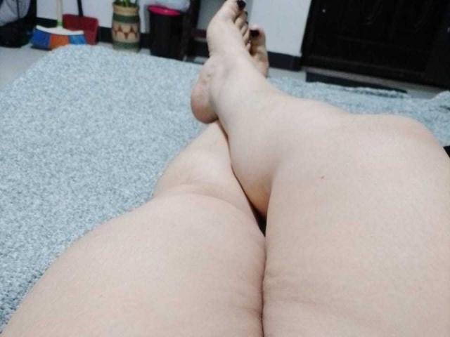 Les jambes de FoxyBell sont croisées aux chevilles, laissant apparaître une peau pâle et des orteils peints en noir. Ses cuisses et ses fesses sont visibles au premier plan, sur un tapis gris..