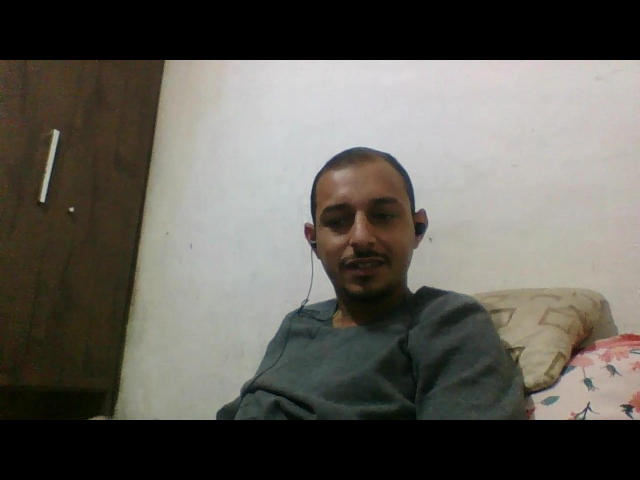PauzudoI has a short mustache, black skin, and a small goatee, wearing a gray sweatshirt, sitting with a slightly annoyed expression, and a pillow behind.