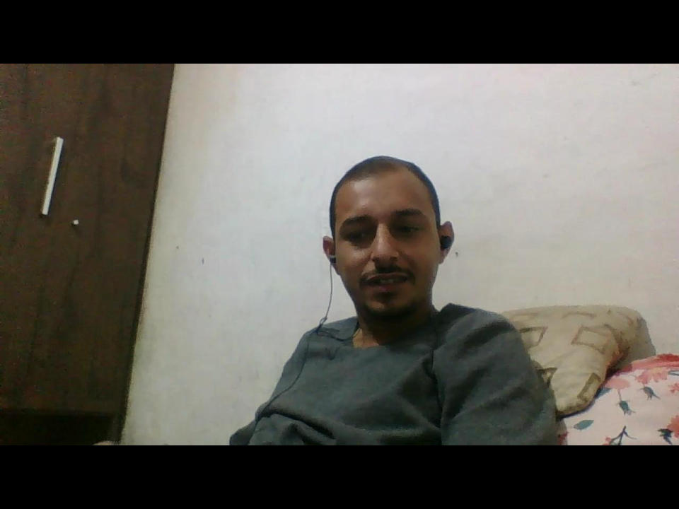 PauzudoI has a short mustache, black skin, and a small goatee, wearing a gray sweatshirt, sitting with a slightly annoyed expression, and a pillow behind.