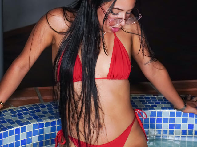 Ciamsth with long black hair, red bikini, small tits, and glasses, leaning on blue-tiled hot tub, showing a tattoo on her right arm. Her pink lips are slightly parted.