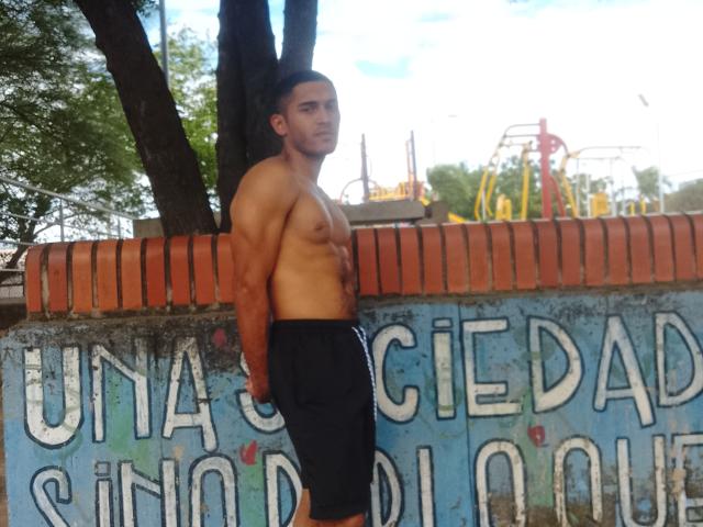 DereckStorm, shirtless, muscular, tan skin, black shorts, standing against a blue and orange graffiti wall, looking at camera, short dark hair, tree in background, outdoor setting.