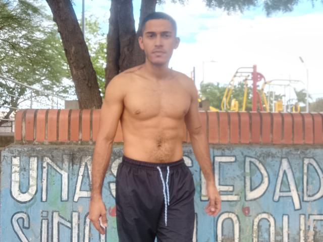 DereckStorm is a shirtless, muscular dude with a hairy chest and black pants standing in front of a brick wall, looking serious. He's got a short, dark haircut.