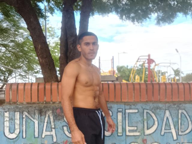 DereckStorm, shirtless with a muscular, tan, hairy chest, wearing black shorts, standing in front of a brick wall, has short dark hair and a serious expression.