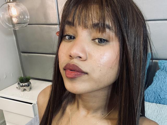 KathaThoms has brown straight hair, bangs, and glossy lips with a slight pout. Her shiny, slightly freckled face shows subtle makeup and a gold necklace.