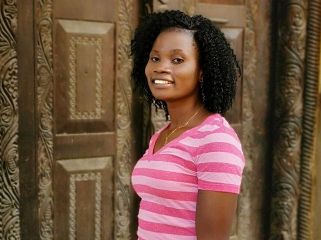 BelaireX, une femme à la peau foncée avec des cheveux noirs bouclés, portant une chemise rayée rose et blanche, souriante, montrant ses dents, debout devant des portes en bois ornées..