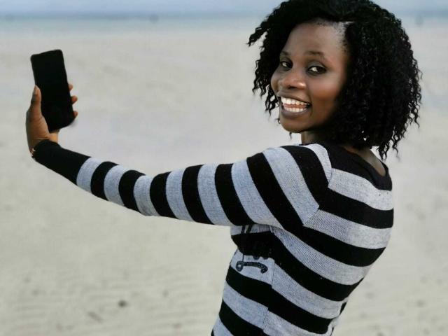 BelaireX, con capelli neri ricci, sorridente, indossa una camicia a righe nere e bianche, tiene in mano uno smartphone nero e sta scattando un selfie su una spiaggia sabbiosa..