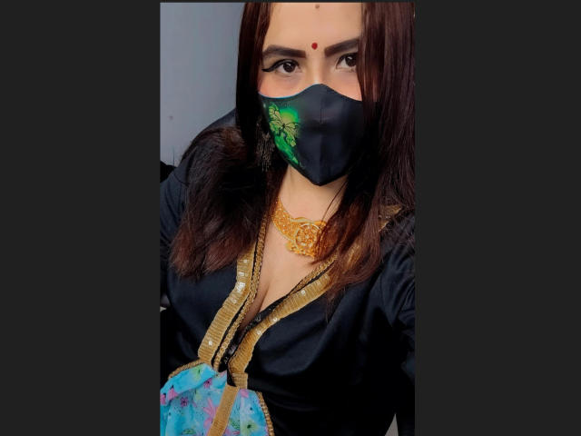 DaliaDubey with dark brown hair, wearing a black mask with green pattern, gold necklace, and black shirt, holding a colorful bag, showing her eyes and a red bindi.