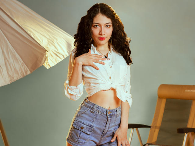 AmeliaTorres with curly dark hair, white buttoned shirt tied at the front, showing her midriff, and blue denim shorts, pointing at her small tits with a soft smile.