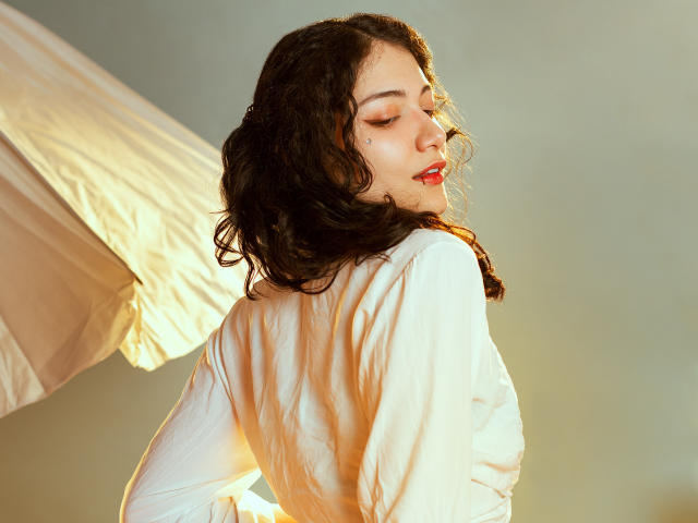 AmeliaTorres with curly dark hair, white shirt, red lipstick, and closed eyes, looking back, soft light on right side, white fabric in background.