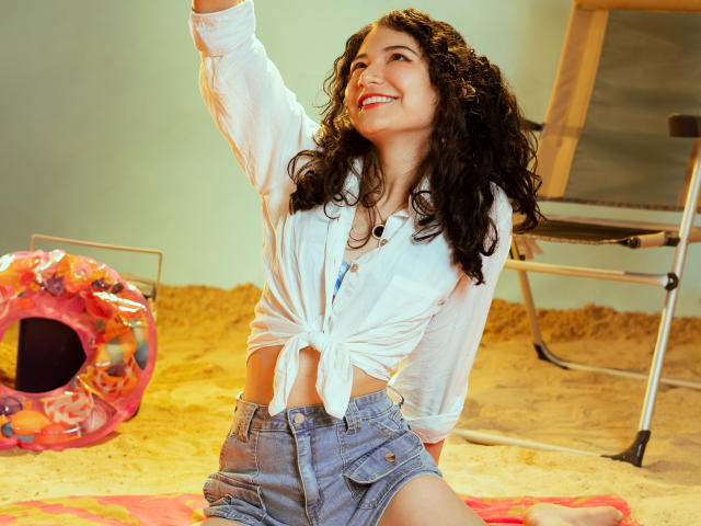 AmeliaTorres with curly black hair, smiling, wearing a tied white shirt and blue denim shorts, raising her arm, showing her cute ass, and red lipstick in a sandy room.