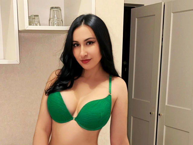 SexyEllene-hot with green bra, showcasing her tits, has long black hair, light skin, and a cute smile, standing in a room with a door and shelf in background.