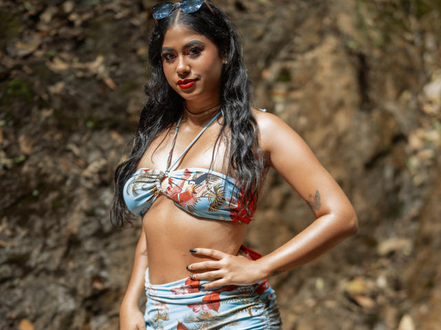 Een vrouw met een gemiddelde borst, gekleed in een bikini met bloemenprint, donker golvend haar, rode lippenstift, een zonnebril op haar hoofd, met haar handen op haar heupen, staand voor een rotsachtige achtergrond..