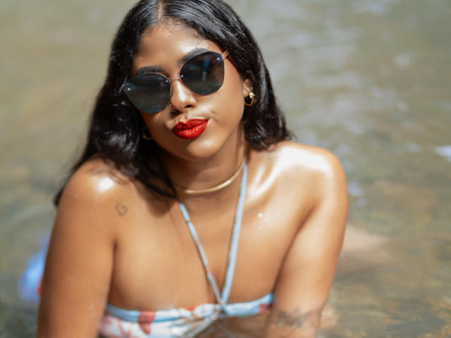 Eine Frau mit dunklem, nassem Haar, die roten Lippenstift und einen blau gestreiften Bikini-Oberteil trägt, sowie eine goldene Halskette. Sie befindet sich im flachen Wasser, blickt nach oben und zieht die Lippen zusammen. Ihre Brüste sind sichtbar..