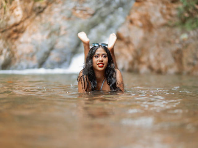 Katika Rouse, met nat donker haar, tilt haar billen op in een rivier, terwijl ze een blauw bikinietje draagt. Haar rode lippen contrasteren met haar bruine huid, en ze heeft een zonnebril op haar hoofd..