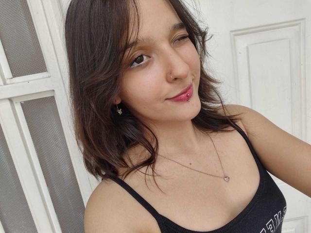 LanaKlaire, con ojos marrones entrecerrados, cabello castaño medio hasta los hombros, un piercing en la nariz, lápiz labial rosa, una camiseta negra, un collar y pequeños aretes, mostrando una ligera sonrisa..