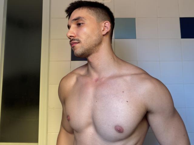 Andosia, shirtless with a muscular build, short dark hair, and a slight beard, showing his bare chest and small, pink nipples. His face is in profile, looking to the side.