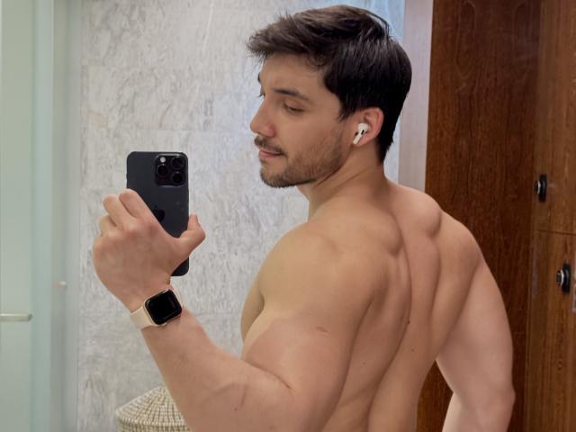 Muscular Andosia with short brown hair, earbuds in, wearing a smartwatch, taking a selfie, showing a strong back and butt, holding a black phone.