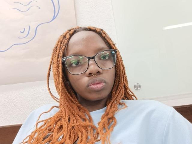 Kastery has dark skin, orange braided hair, green glasses, a nose ring, and a light blue shirt, with puffy lips. She's looking straight at the camera.