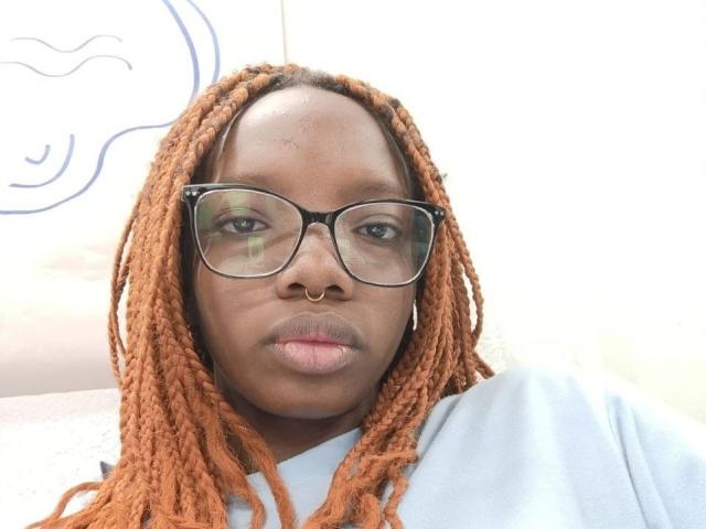 Kastery has dark brown skin, orange braided hair, black-framed glasses, a gold nose ring, and a serious expression, wearing a light blue shirt.