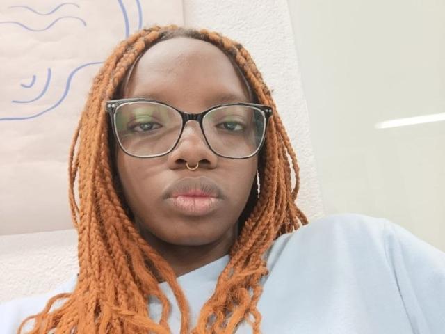 Kastery with orange dreadlocks, black glasses, nose ring, and light blue shirt, pouting slightly, green eyeshadow, white wall in background.