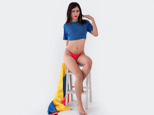 TamaraGarcia with medium tits, red panties, and blue crop top, sitting on a stool, yellow and blue flag on the ground, white background, bare feet, red lipstick.