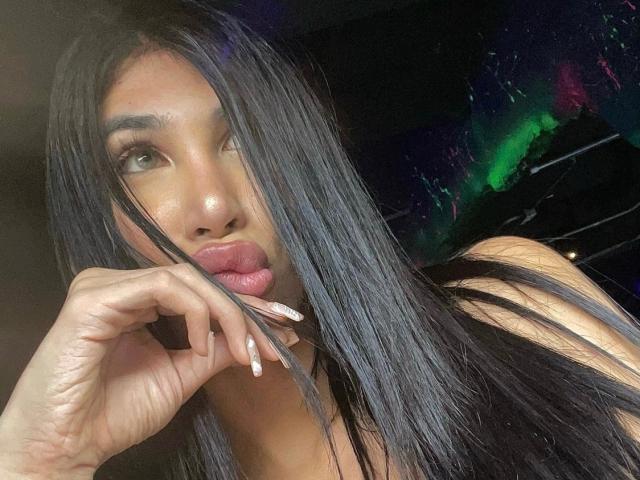AndreaBigCock, avec ses longs cheveux noirs, ses yeux marron et sa peau brillante, faisant la moue avec la langue dehors, touchant ses lèvres avec des ongles manucurés, dans un selfie sexy et confiant..