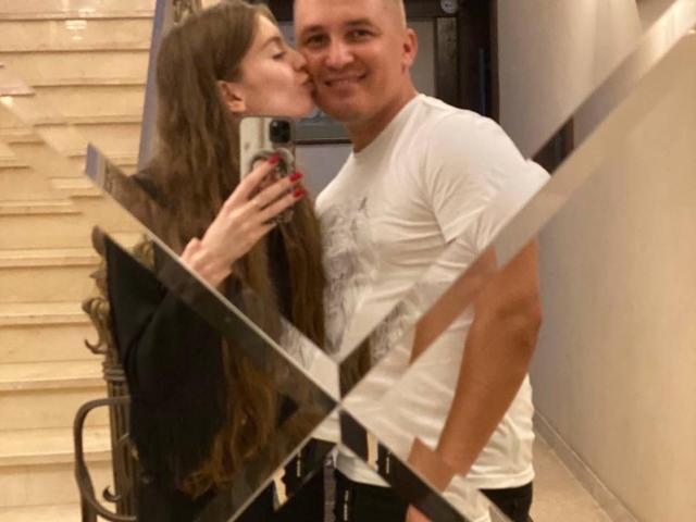 LoganEmma with long brown hair kissing a bald man wearing a white shirt. She's holding a phone with red nails, standing next to a glass railing. His hand's in his pocket.