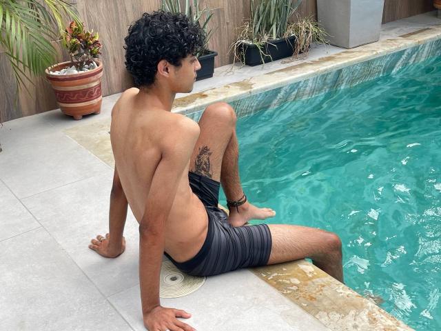 WorldSexxxADS with curly black hair, tan skin, and a tattoo on his right thigh, sitting by a pool, wearing black swim trunks, showing his butt, with legs in water.