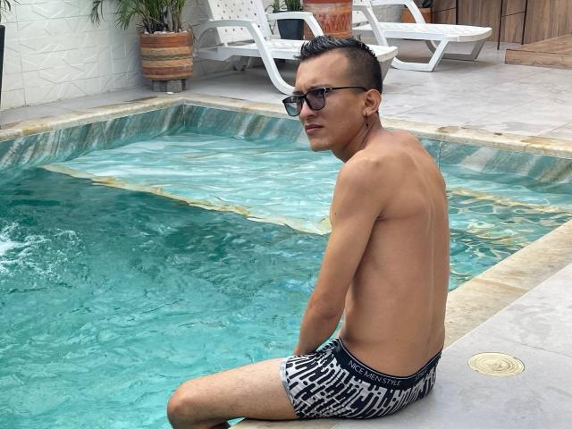 WorldSexxxADS with short black hair, sunglasses, and a slim build, sitting in a pool wearing black and white patterned swim trunks, showing his ass, looking back.
