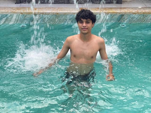 Shirtless WorldSexxxADS with curly black hair, standing in a pool, water splashing around, small tits, and a visible ass, wearing green swim trunks, smiling.
