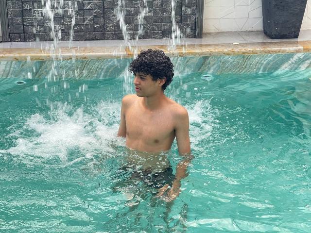Shirtless WorldSexxxADS with curly black hair stands in a turquoise pool, water splashing around his bare ass, looking to the side with a slight smile.