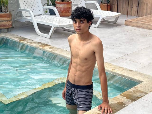WorldSexxxADS, slim with curly black hair, tan skin, wearing black and gray striped swim trunks, stands in a pool, showing a toned, flat chest, and a visible, flaccid, small penis.