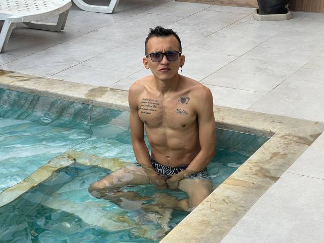 WorldSexxxADS, shirtless, sitting in a small pool, wearing zebra-striped swim trunks, tattoos on chest, short black hair, sunglasses, light skin, medium-sized ass, looking at camera.