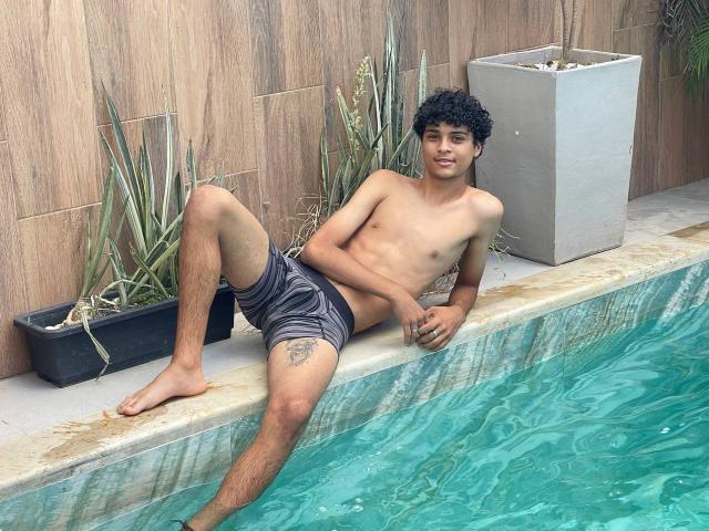 WorldSexxxADS, shirtless, with curly black hair, and gray shorts, leans on a pool's edge, legs in water, showing a tattoo on his right thigh, smiling.