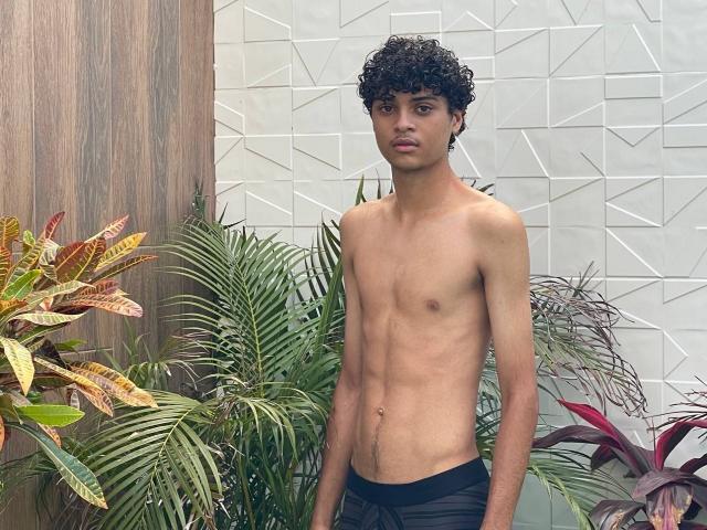 Shirtless WorldSexxxADS with curly black hair, slim build, and small ass, wearing black pants, standing in front of green plants and a geometric white wall.