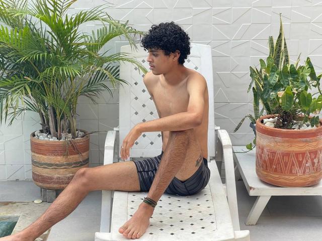 WorldSexxxADS with curly black hair, shirtless, wearing gray shorts, sitting on white chair, legs crossed, tattoo on right thigh, black bracelet, two potted plants, textured gray wall.