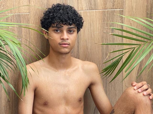 Shirtless WorldSexxxADS with curly black hair, brown skin, and a toned chest, sitting between green palm leaves, looking straight at the camera with a neutral expression.