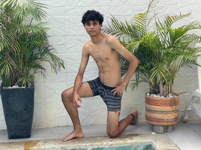 WorldSexxxADS, slim with curly black hair, tan skin, wearing striped black and gray shorts, kneeling by pool, green potted plants, white textured wall, barefoot, looking at camera.