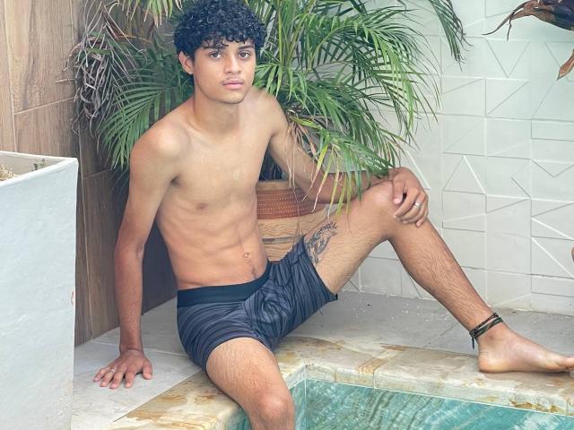 WorldSexxxADS, shirtless, curly black hair, tan skin, black shorts, sitting by a pool, tattoo on right thigh, ankle bracelet, pensive expression, green plant in background.