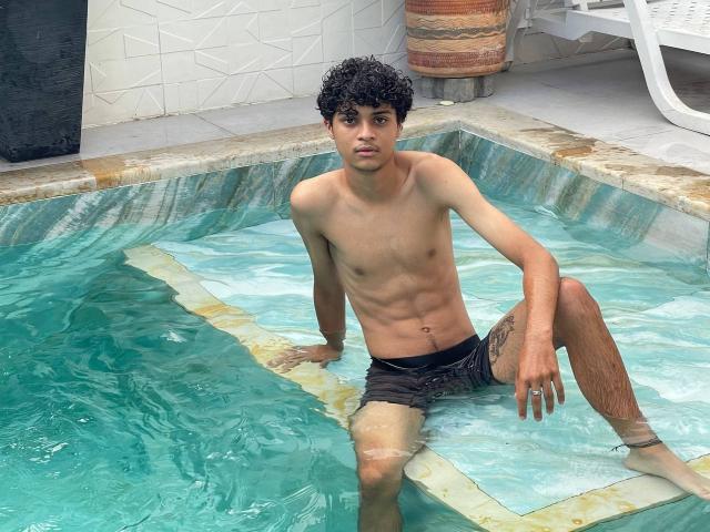 WorldSexxxADS is a skinny, young, dark-haired guy with a tattoo on his thigh, sitting in a pool wearing black swim trunks, showing his small, bare ass and cock.