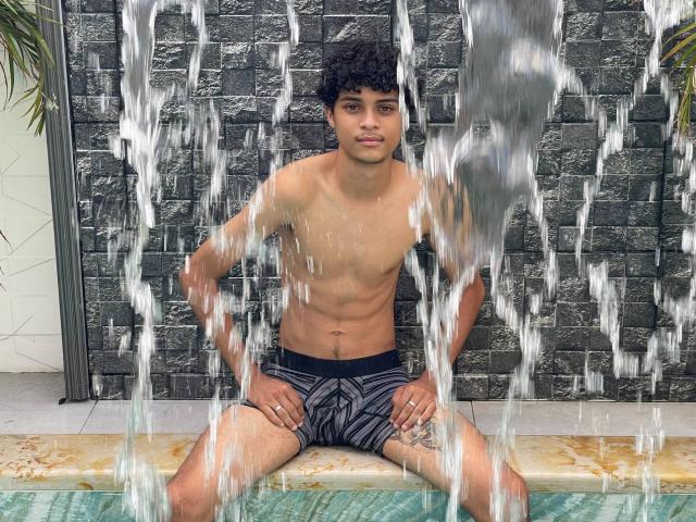 WorldSexxxADS, a curly-haired, tan-skinned dude with a toned, flat-chested body, sits in a pool, wearing black and gray striped swim trunks, with water spraying in front.