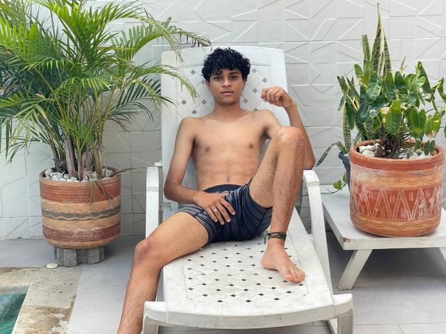 WorldSexxxADS with curly black hair, slim, shirtless, wearing dark shorts, ankle bracelet, leg raised on white chair, surrounded by potted plants, white tiled wall, and a pool edge.