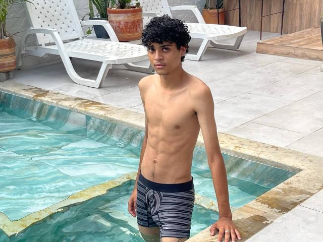 WorldSexxxADS with curly black hair, tan skin, and a slim, toned, flat-ass body, wearing black and gray striped swim trunks, standing in a pool, looking at the camera.