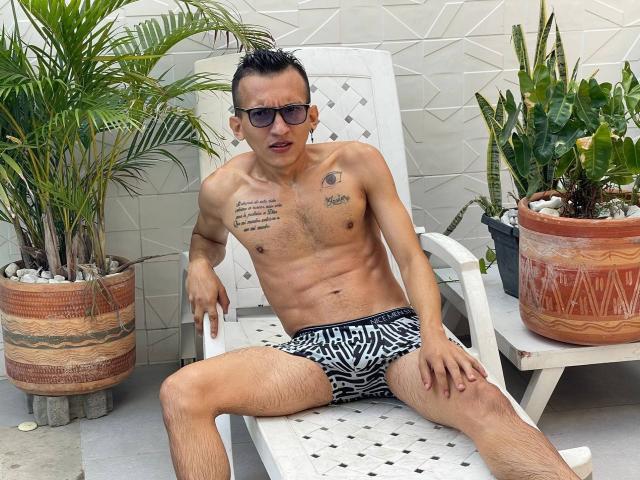 WorldSexxxADS, shirtless, wearing zebra-print boxer briefs, sitting on a white lounge chair, legs spread, tattoos on chest, short dark hair, sunglasses, potted plants, light skin, muscular.
