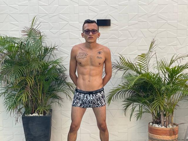 WorldSexxxADS, shirtless, wearing black and white patterned shorts, standing with hands behind back, tattoos on chest, short dark hair, sunglasses, potted plants on either side.