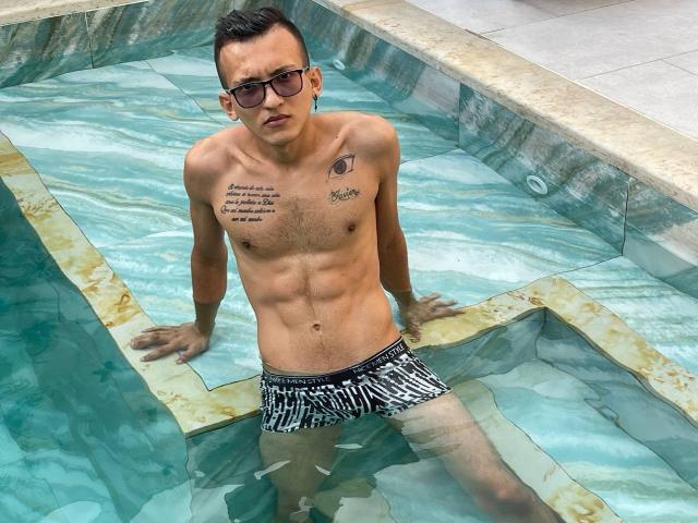 WorldSexxxADS, shirtless, in a pool, wearing black and white striped swim trunks, tattoos on chest, dark sunglasses, short dark hair, muscular, standing with hands on pool edge.